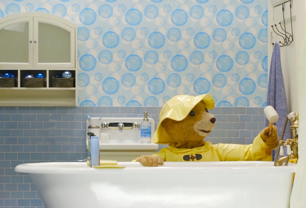 Paddington Bear dressed in a yellow raincoat and hat in the bathroom at the neighbors house getting ready for Christmas in Paddington Saves Christmas at Seattle Children's Theatre 2022